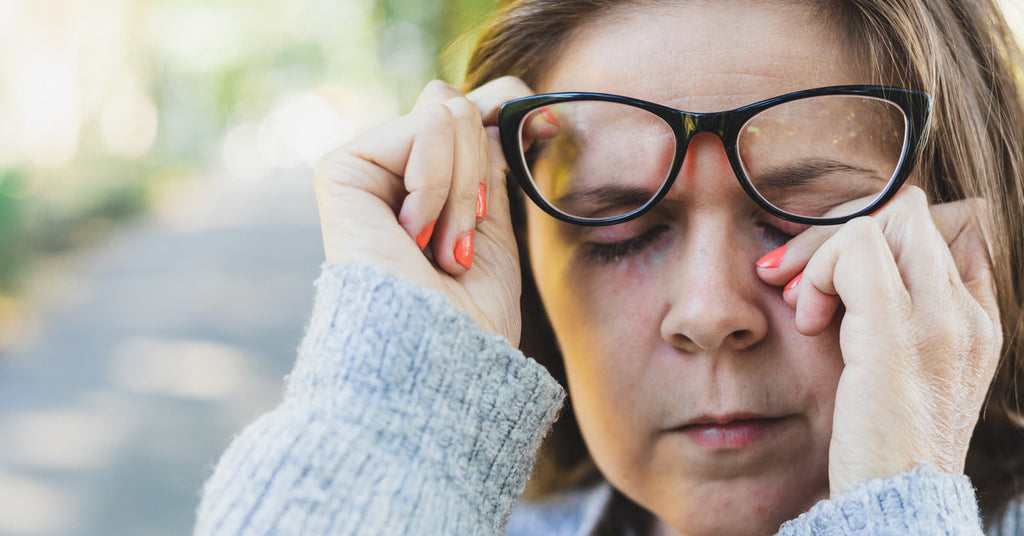 A close-up of a woman standing outside and lifting up her glasses with one hand while rubbing her eye with the other.