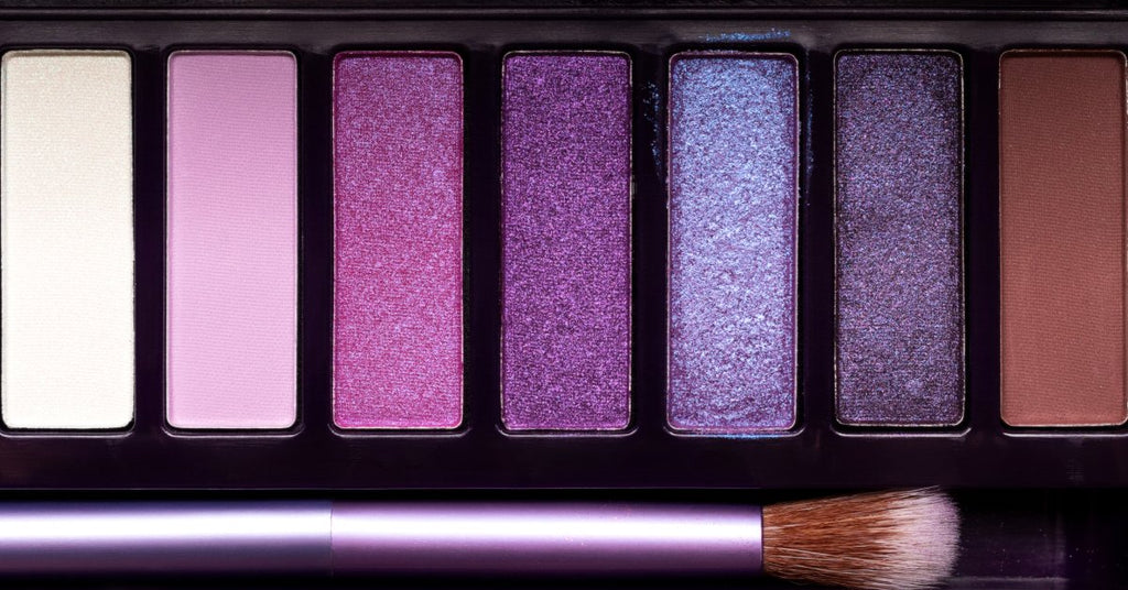 An eye shadow palette containing a brush, a highlight color, a contour, and five vibrant, glittery shades of pink and purple.