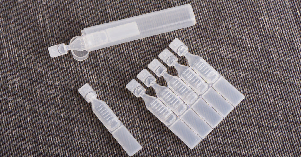 Seven containers of artificial tears are sitting on a grey fabric and one container is in a larger plastic bottle.