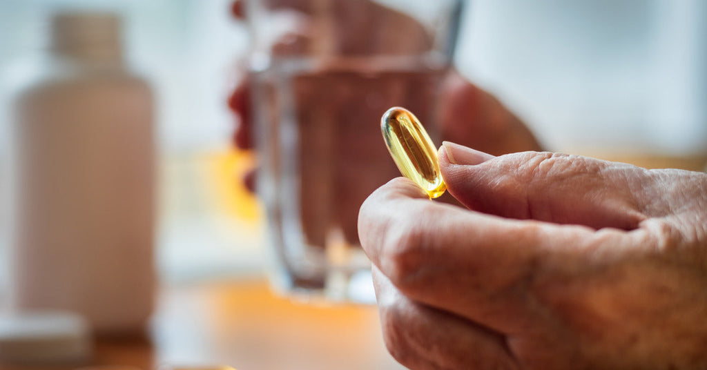 A person is holding an Omega-3 supplement in their left hand and a full cup of water in their right hand.